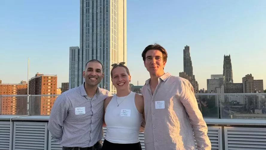 Three people wearing name badges pose together on a rooftop terrace at sunset, with tall city buildings and a clear blue sky in the background.
