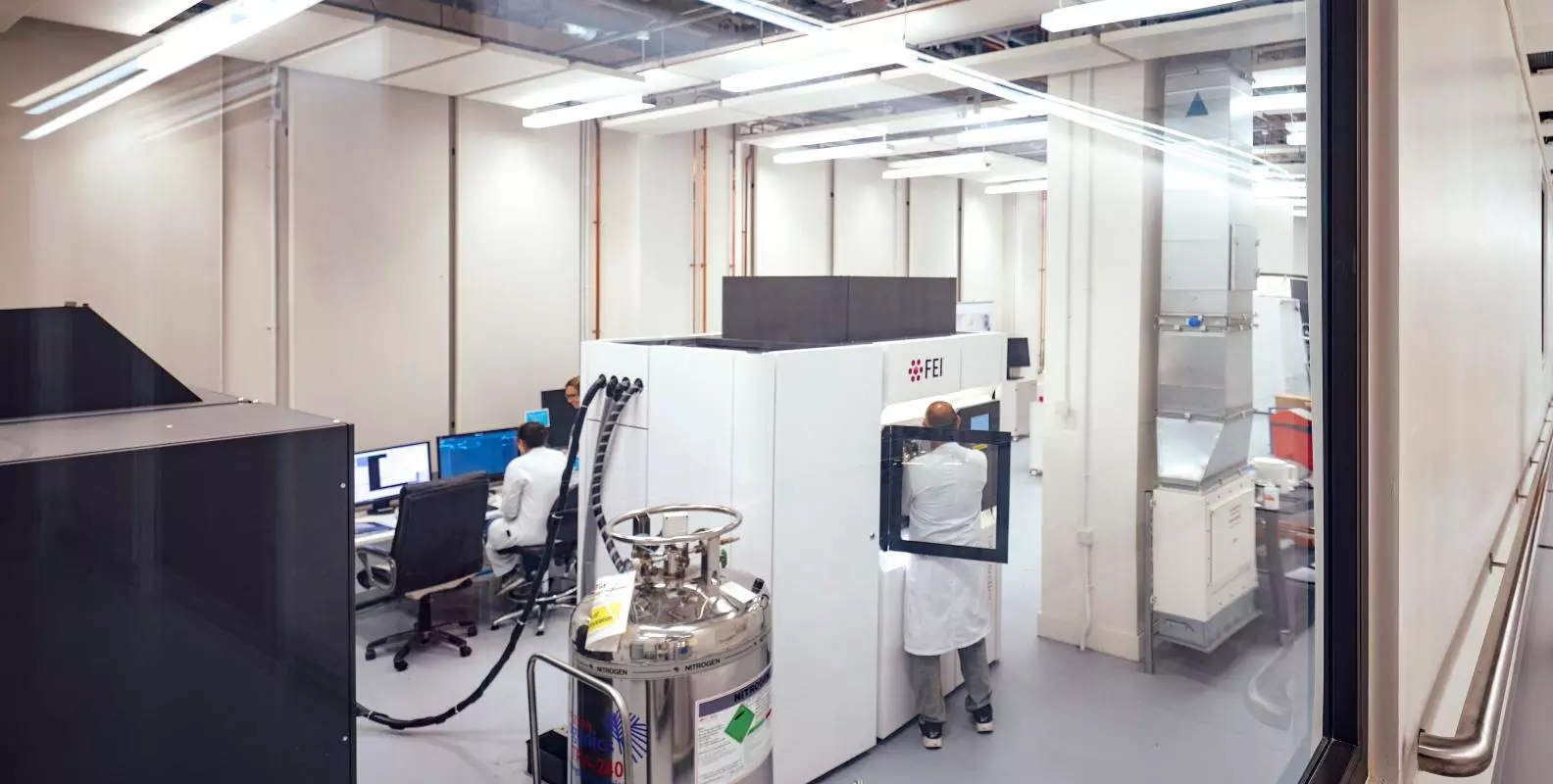A view through glass into a specialist microscopy laboratory, with a large cryo-EM, two researchers in lab coats and metal liquid nitrogen storage tanks.
