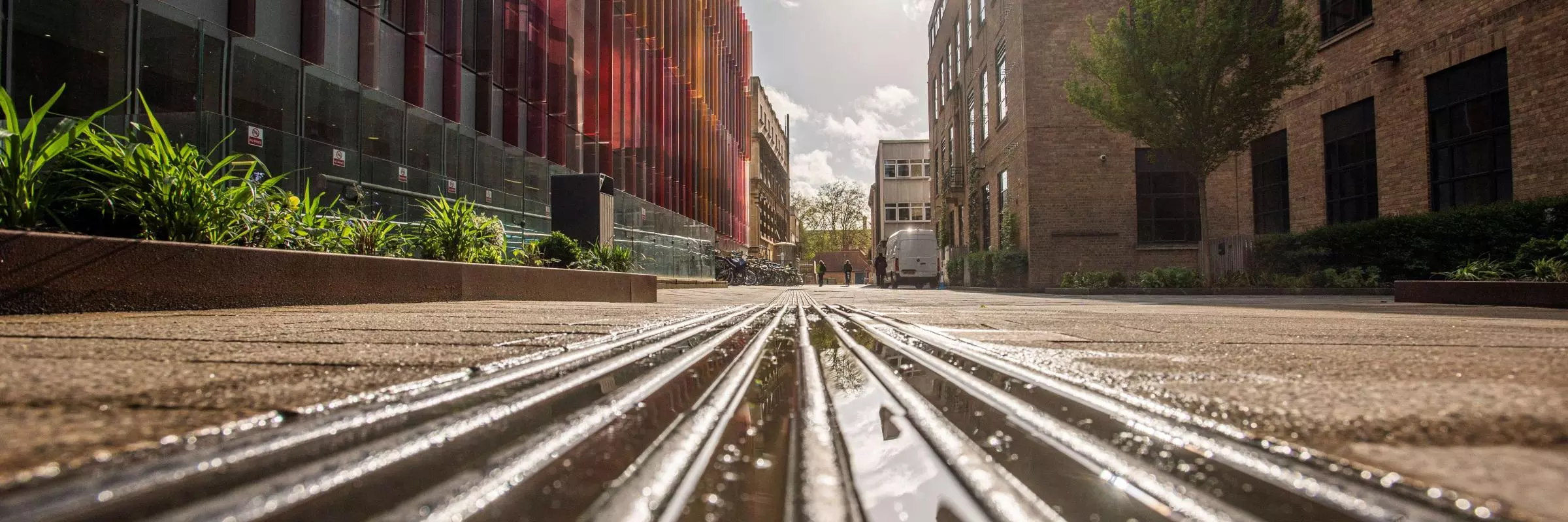 Low-angle view of rain-soaked metal tracks running between modern buildings, reflecting sunlight beneath a partly cloudy sky.